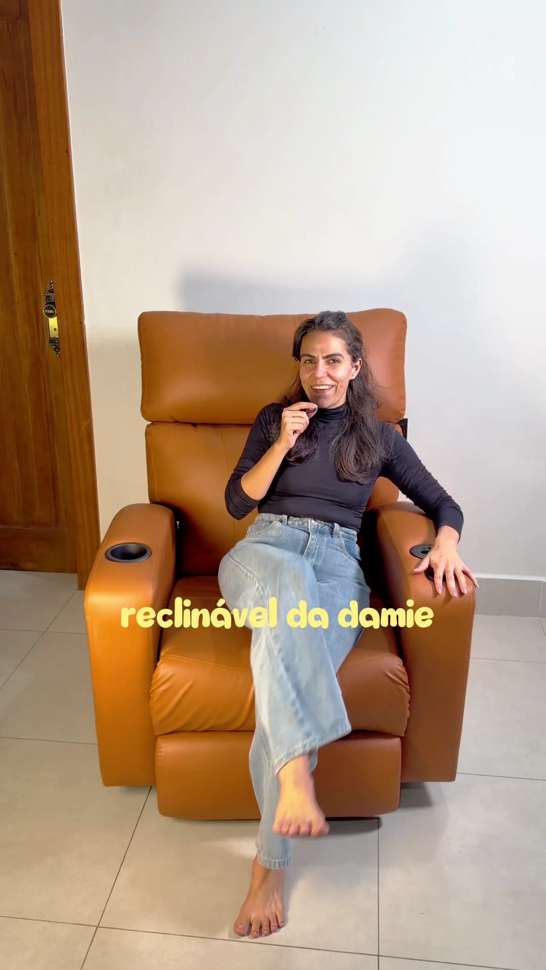 Cecília sentada na Poltrona Reclinável Damie 2.0 em couro caramelo, na sala de casa.